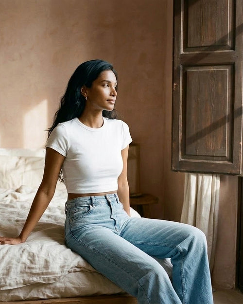 Woman wearing the Hallberd Cotton Crop Top in white with light-wash wide-leg jeans, sitting on a linen bed by wooden shutters in morning light