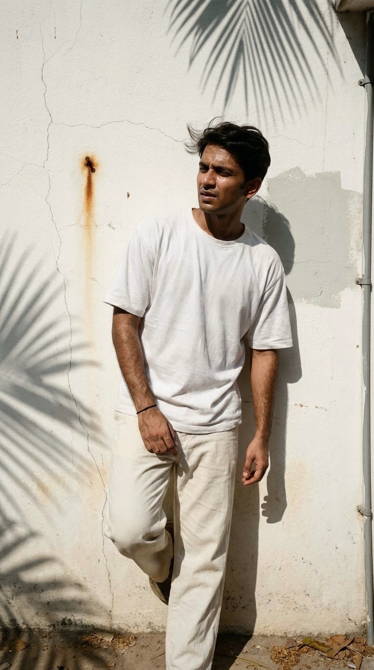Man wearing the Hallberd Oversized Plain T-Shirt in white with ecru linen trousers, leaning on a sunlit clay-pink wall in a courtyard with an olive tree