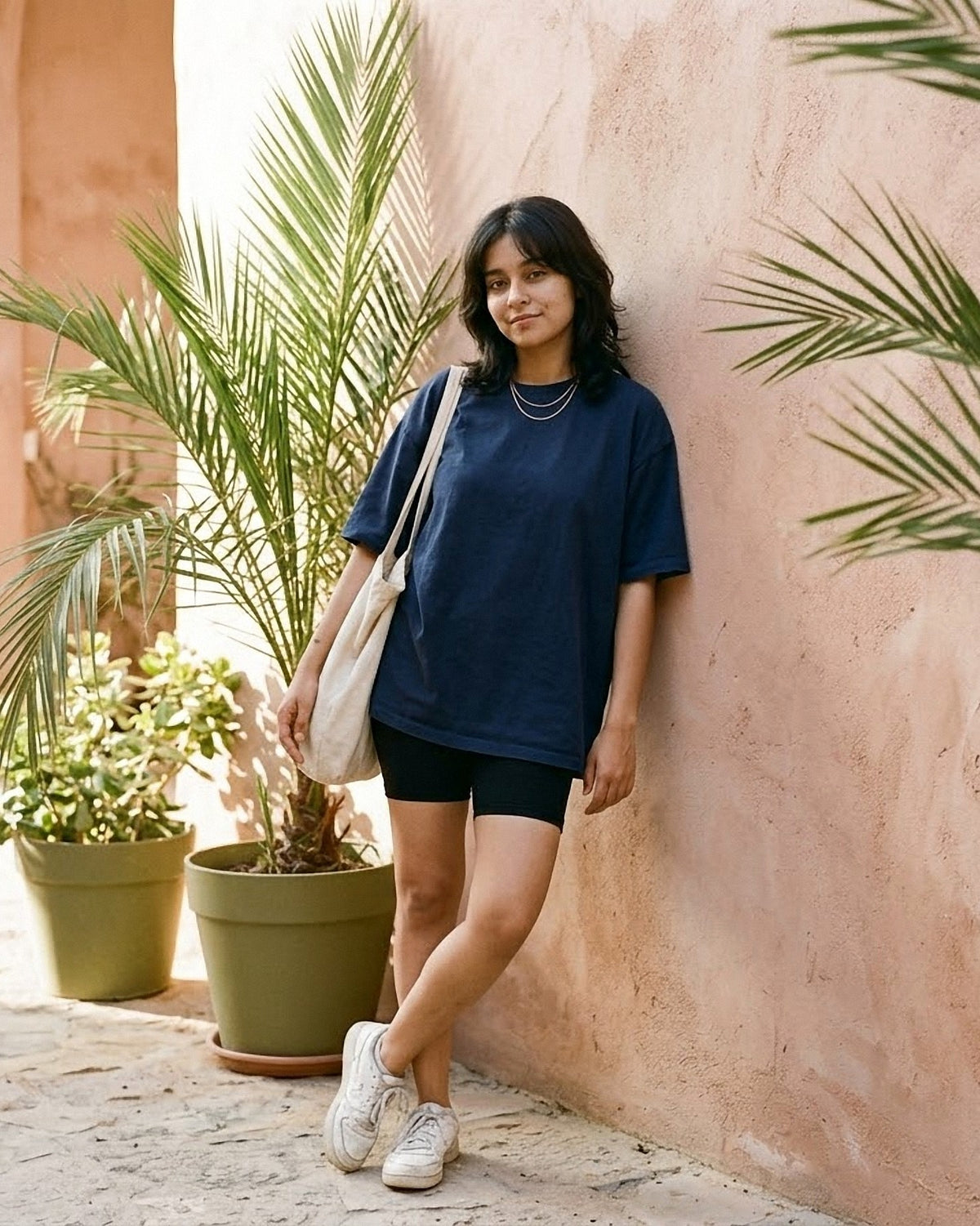 Woman wearing the Hallberd Women's Oversized T-Shirt in navy with black bike shorts and white sneakers, leaning on a clay-pink wall beside potted palms
